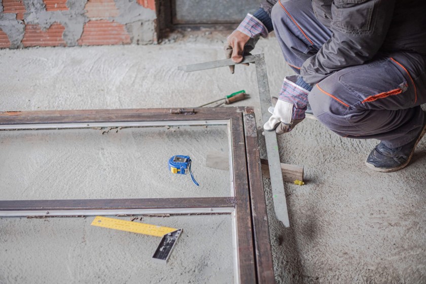 Worker measuring and aligning a metal frame on a concrete floor using a square tool and tape measure.