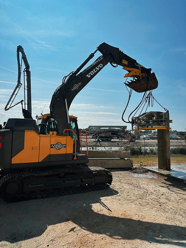 Volvo excavator with a heavy-duty attachment lifting a large metal structure at a construction site under a clear blue sky.