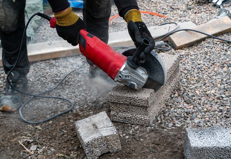 A worker using a red angle grinder to cut concrete blocks on a gravel surface in Texas & Louisiana